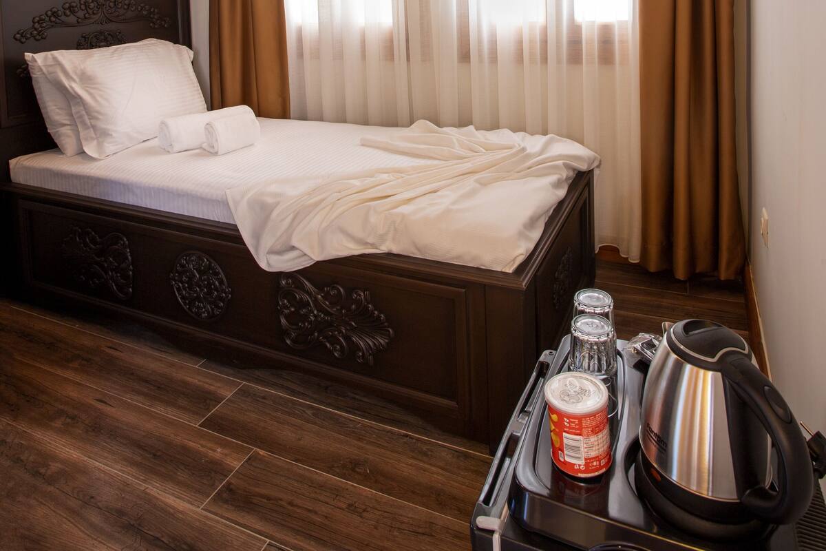A hotel room with a wooden ceiling, a bed with white linens, a window with sheer curtains, and a kitchenette area with a kettle and a can of drink.