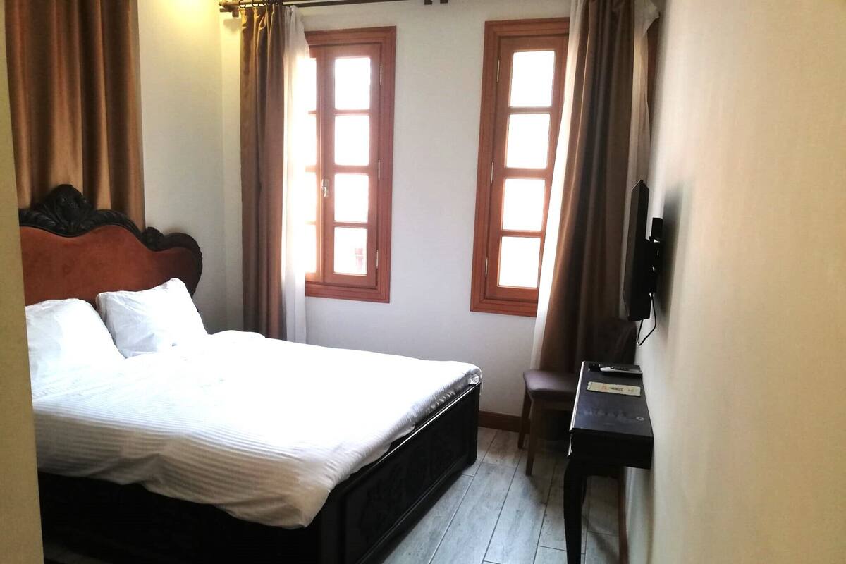 A bedroom with a bed, two windows, a desk, and a television.