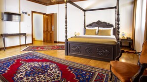 A four-poster bed with a dark wooden frame and gold accents, a red and blue patterned rug, a wooden floor, a wooden desk, a television, and a door in the background. -  WOO Town  (Antalya)