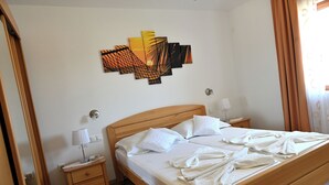 Una habitación de hotel con una cama de madera, ropa de cama blanca, una mesita de noche con lámpara, una obra de arte en la pared y una ventana con cortinas.