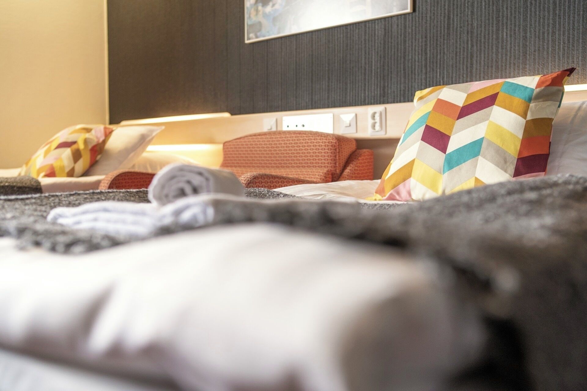 A hotel room with a bed, pillows, a blanket, and a wall-mounted light switch.