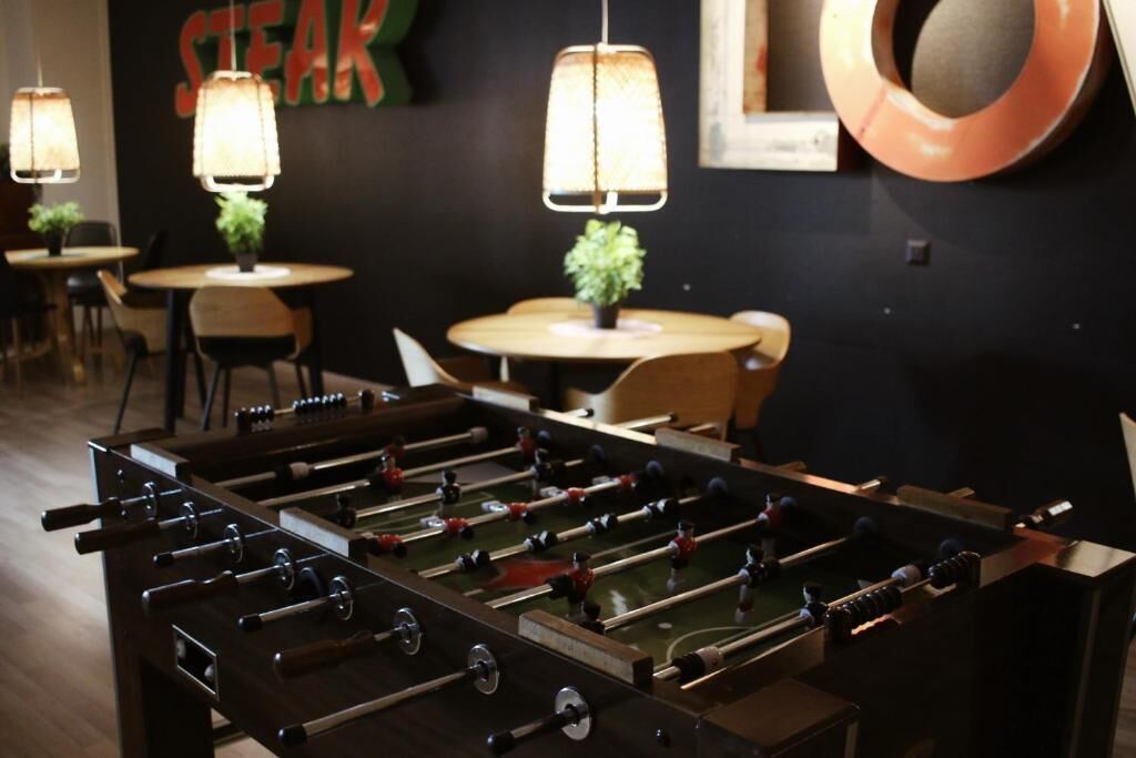 Una habitación con una mesa de fútbol de mesa, mesas con sillas, lámparas colgantes y una pared con un letrero.