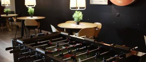Una habitación con una mesa de fútbol de mesa, mesas con sillas, lámparas colgantes y una pared con un letrero.