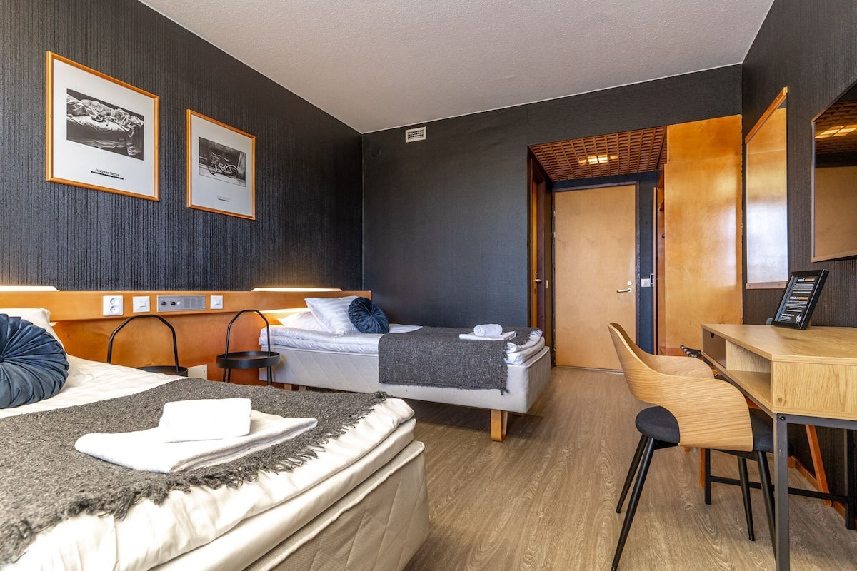 A hotel room with a bed, a desk, a chair, and two framed pictures on the wall.