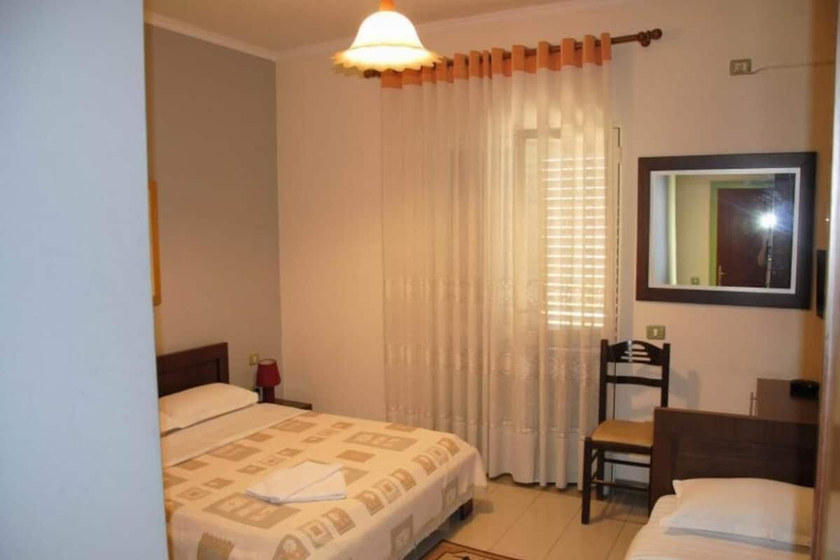 A bedroom with a bed, a chair, a desk, a mirror, and a window with curtains.