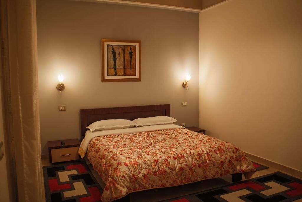 A bedroom with a bed, two wall-mounted lights, a painting, and a geometric patterned rug.