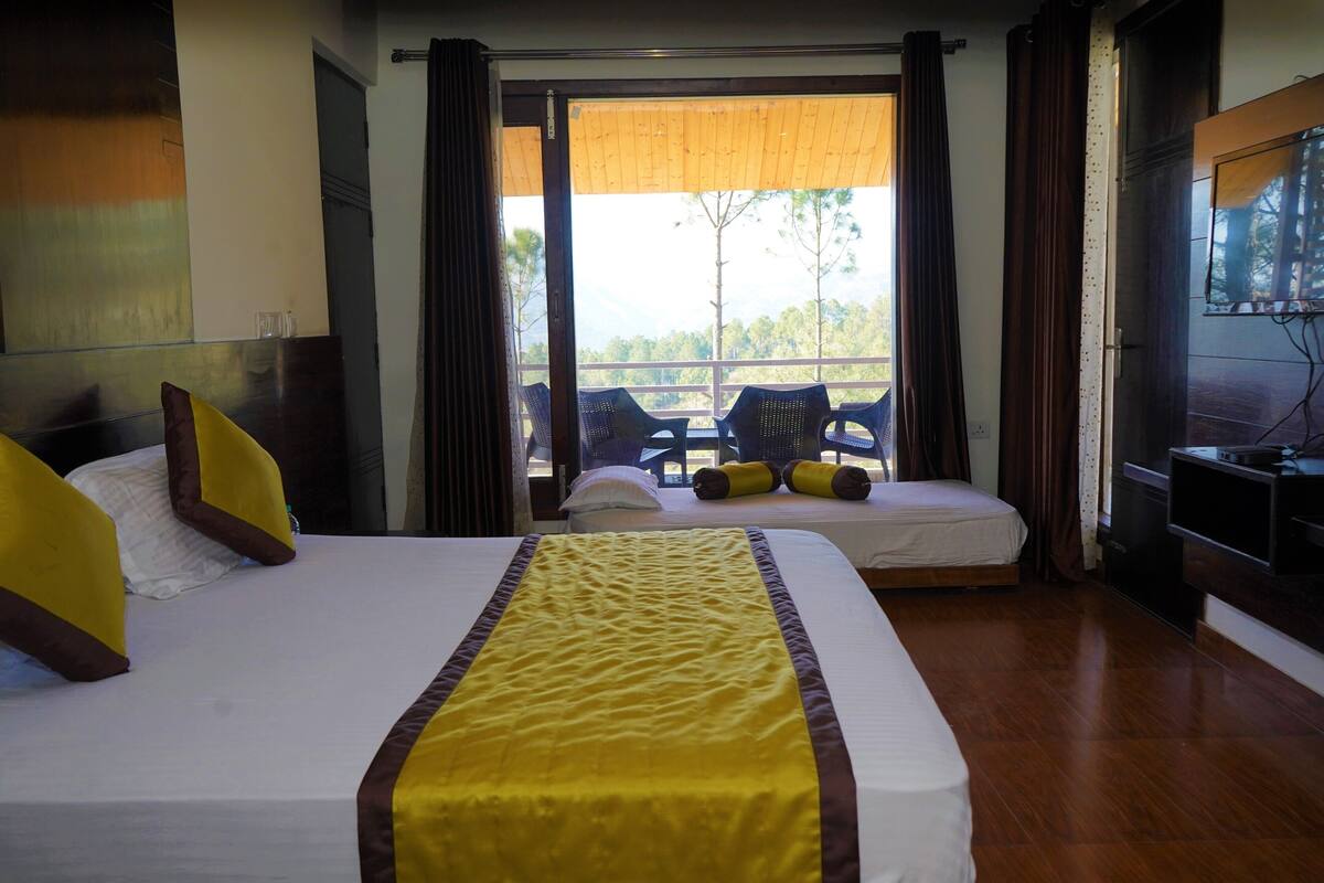 Habitación de hotel con una cama grande, sofá cama, televisión y balcón con vistas a árboles y montañas.