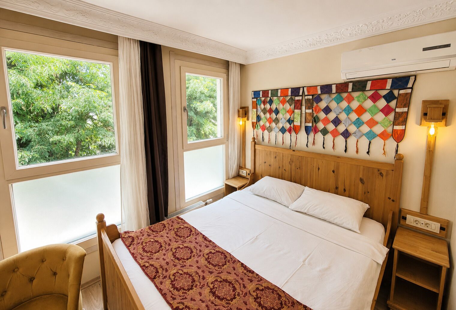 A hotel room with a large bed, a wooden headboard, a window with curtains, a bedside table with a lamp, and a wall-mounted air conditioning unit.