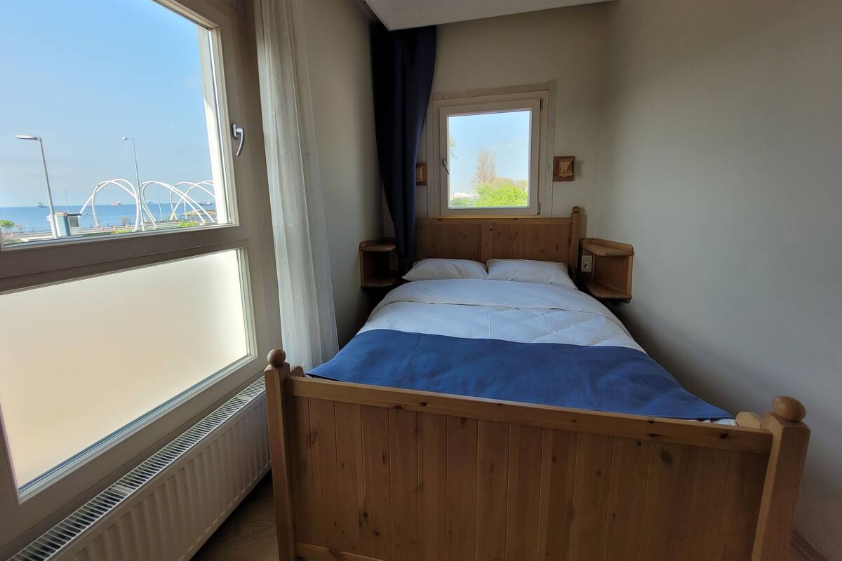 A bedroom with a wooden bed, a window with curtains, a radiator, and a view of a bridge and greenery.