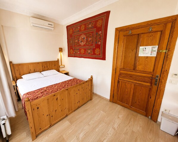 A wooden bed with a headboard, a bedside table, a wall-mounted air conditioner, a red patterned tapestry, and a wooden door with a lock.