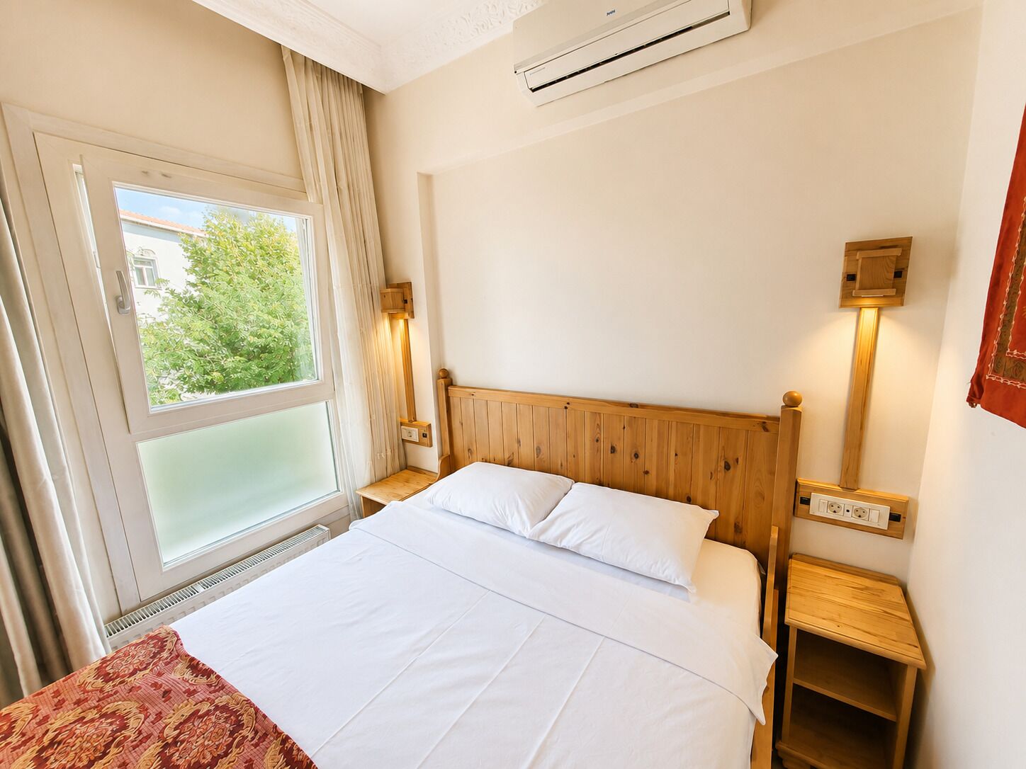 A hotel room with a bed, a window with curtains, an air conditioning unit, and a bedside table with a lamp.
