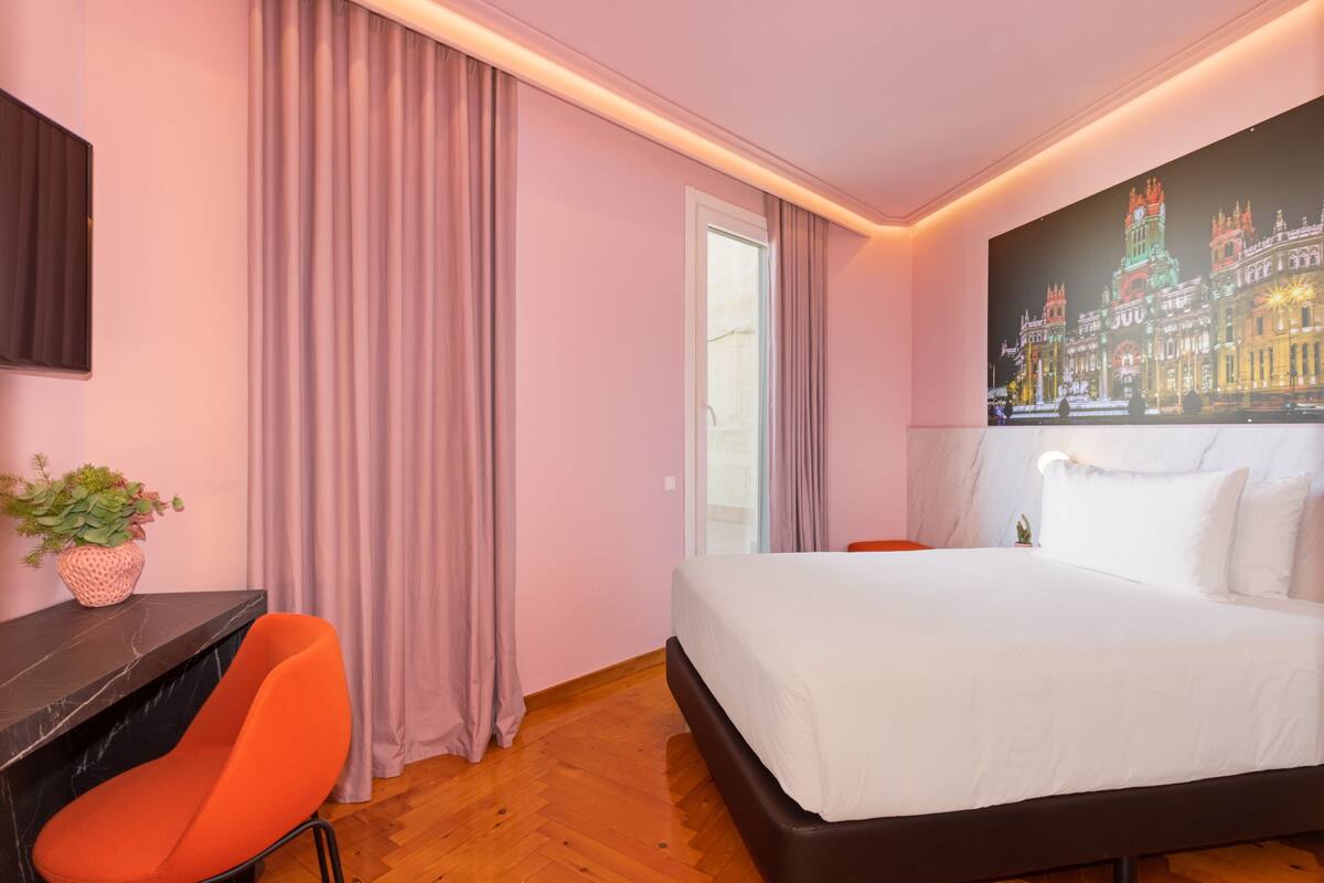 A modern hotel room with a large bed, a desk with a chair, a potted plant, and a large window with curtains.