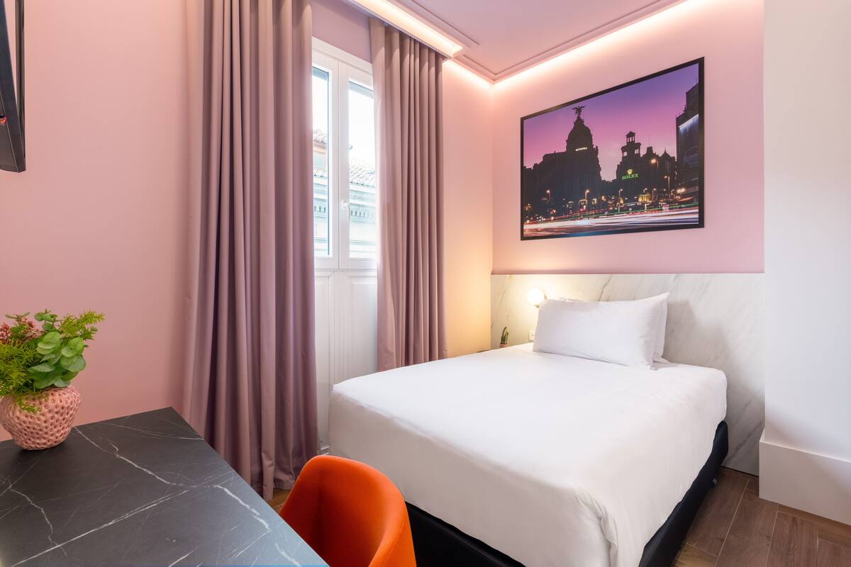 A hotel room with a bed, a desk, a chair, a potted plant, and a cityscape painting on the wall.