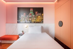 Ein Hotelzimmer mit einem großen Bett, einem Nachttisch, einem kleinen Tisch mit einer Pflanze und einem Stadtbild an der Wand.