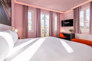 Ein Hotelzimmer mit einem großen Bett, einem an der Wand befestigten Fernseher, einem Schreibtisch mit Stuhl und Blick auf Gebäude durch die Fenster.