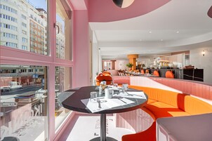 Eine moderne Hotellobby mit einem runden Esstisch, einer Sitzecke mit orangefarbenen Kissen und Blick auf die Stadt durch große Fenster.