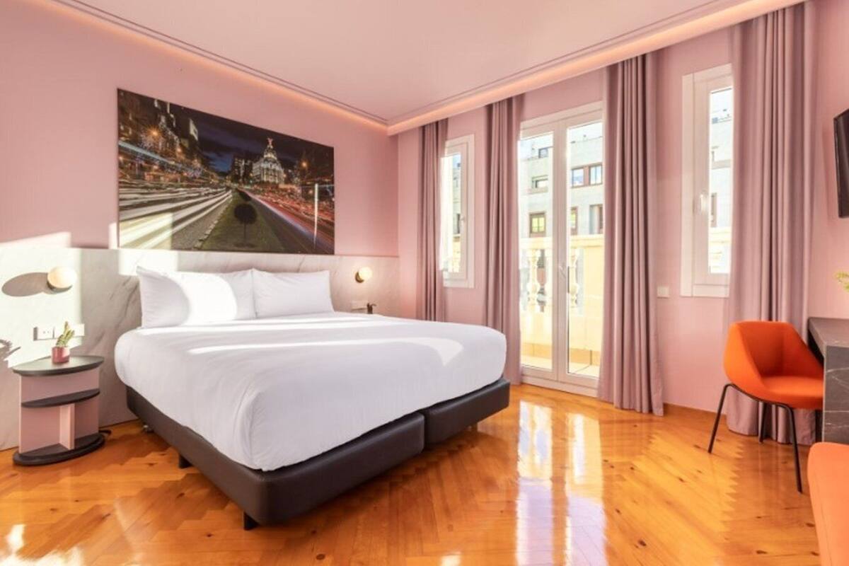 A modern hotel room with a large bed, a night cityscape painting, a balcony with curtains, and a small orange chair.