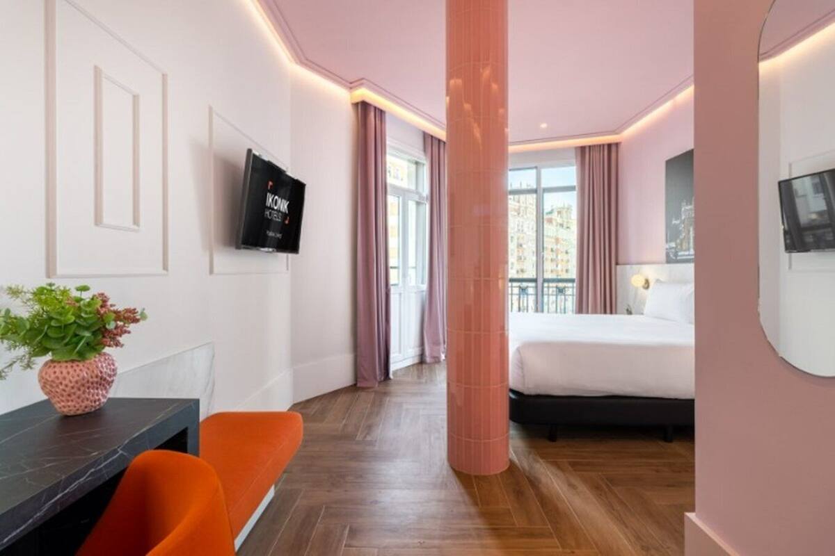 A modern hotel room with a bed, a TV, a desk with a chair, and a potted plant.