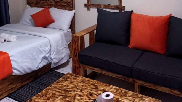 A hotel room with a bed, a sofa, a wooden headboard, a mirror, and a small table with a roll of toilet paper.