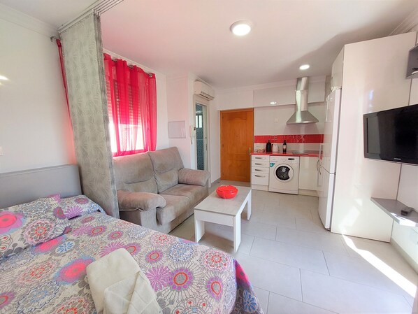 A compact hotel room with a bed, a sofa, a small table, a washing machine, a TV, and a kitchenette.