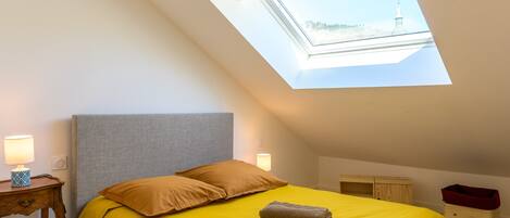 A bedroom with a skylight, a bed with yellow bedding, a bedside table with a lamp, and a wooden dresser.