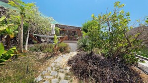 Edificio de piedra con un sendero de jardín que lo conduce, rodeado de plantas tropicales y una hamaca.