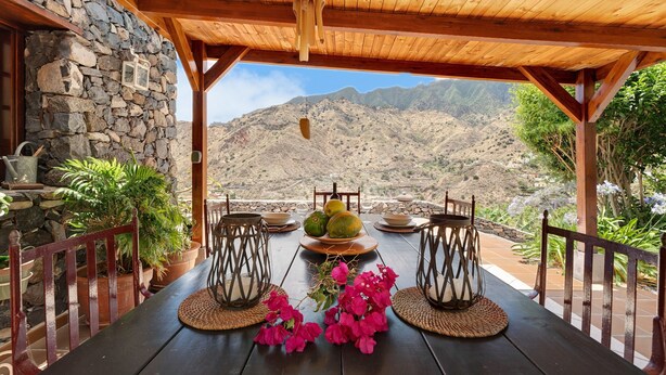Una mesa de madera dispuesta con frutero, velas y flores, con vistas a un paisaje montañoso.