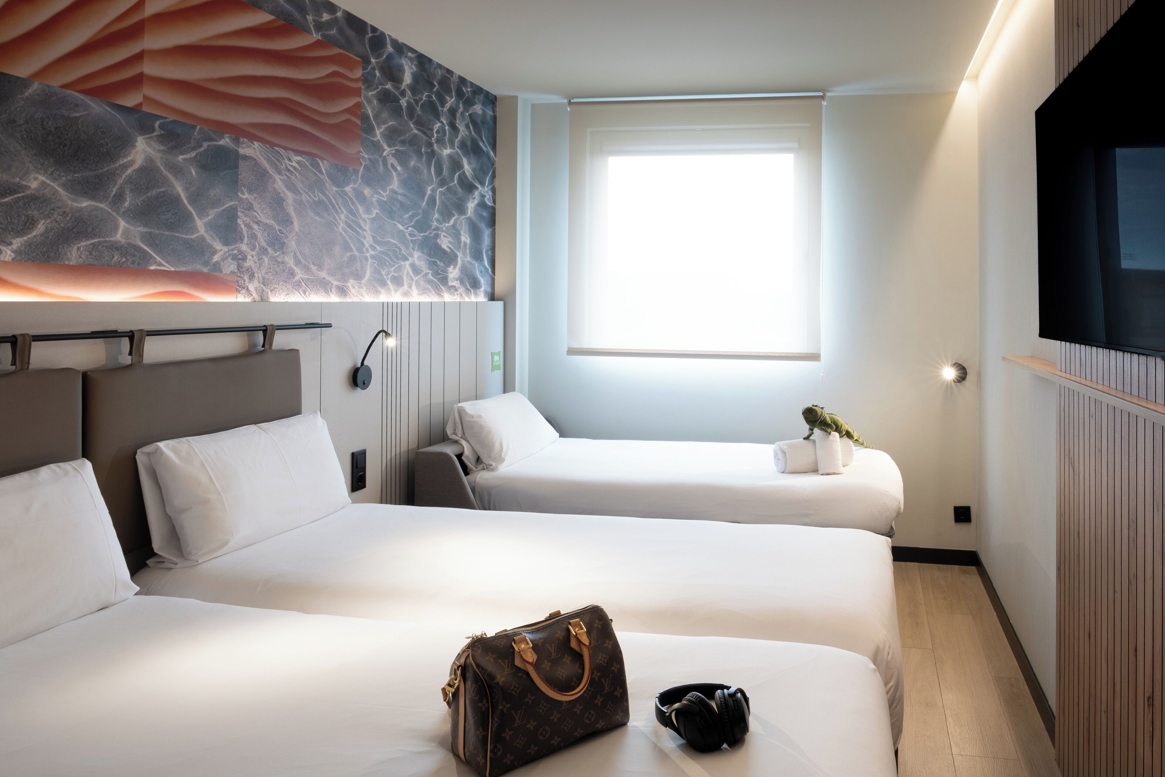 A hotel room with two beds, a large window, a flat-screen TV, and a wall with a textured design.