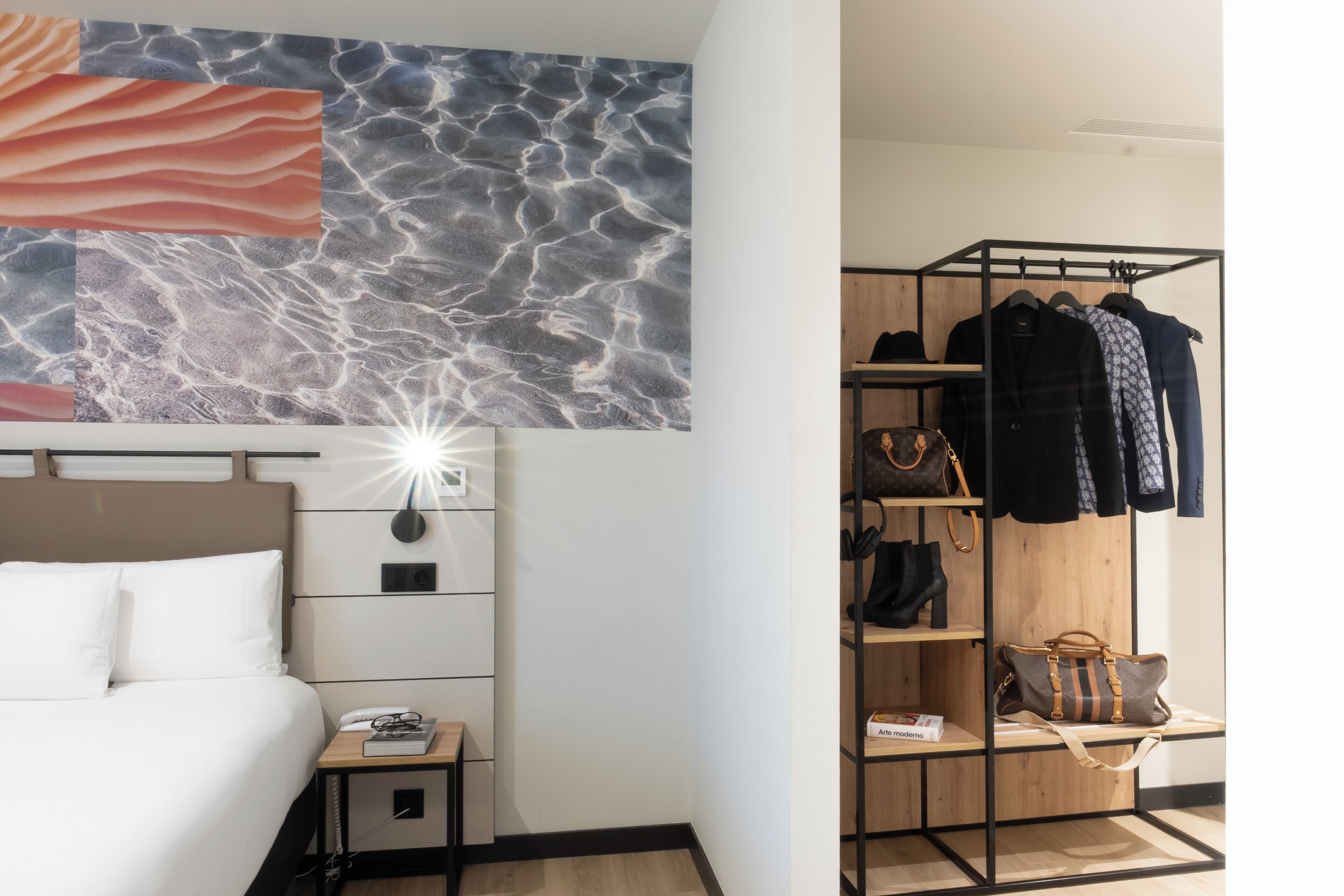Habitación de hotel con cama, mesita de noche, decoración en la pared y un perchero con bolsas.