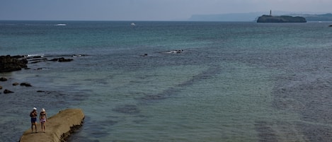 A coastal scene with a concrete pier extending into the water, two people standing at the end, and a lighthouse in the distance.