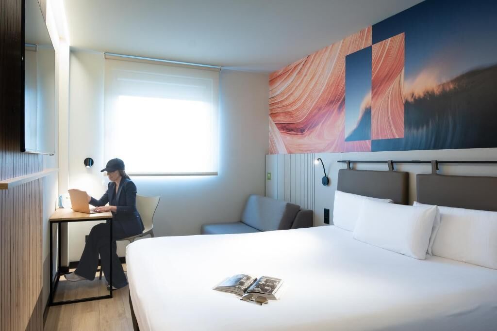 A hotel room with a bed, a desk with a laptop, a window, and a large abstract painting on the wall.