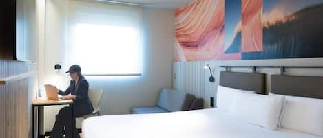 A hotel room with a bed, a desk with a laptop, a window, and a large abstract painting on the wall.