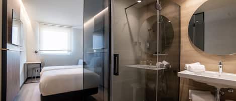 Una habitación de hotel moderna con ducha de vidrio, una cama grande, una mesita de noche, un escritorio pequeño y un espejo amplio.