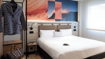A hotel room with a large bed, a nightstand, a lamp, a painting on the wall, a hanging garment, and a suitcase.