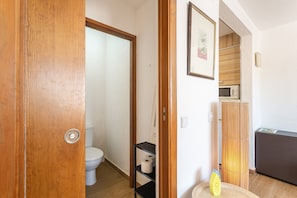 Et badeværelse med toilet, en hylde med toiletpapir, et billede i ramme på væggen og en mikroovn i det tilstødende rum.
