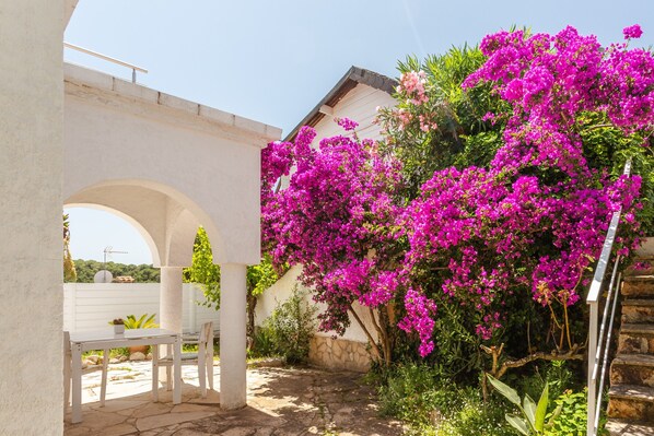 A white outdoor seating area with a stone floor, surrounded by lush greenery and vibrant pink bougainvillea flowers.