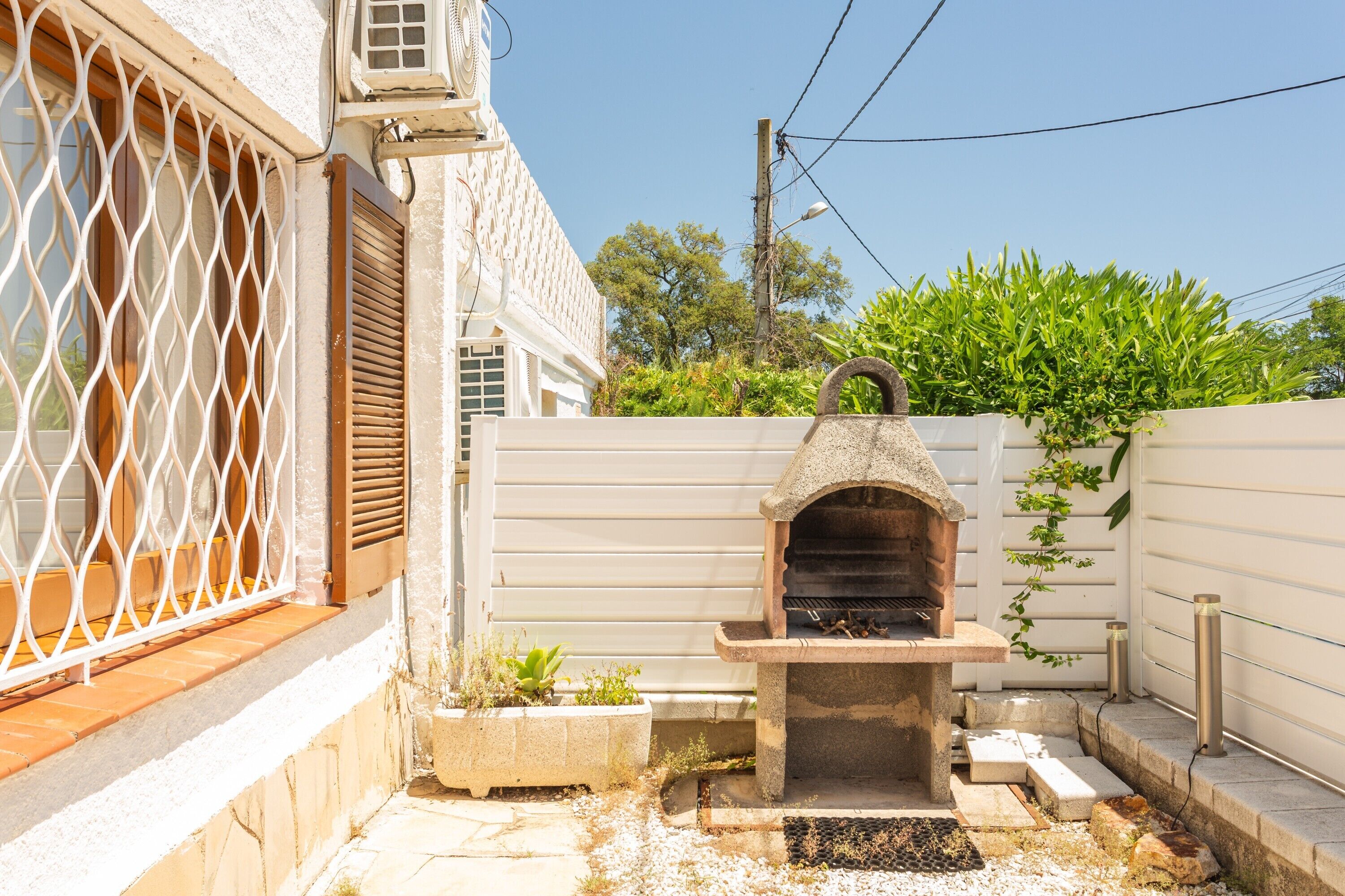 Una parrilla de piedra al aire libre, un edificio blanco con ventana de celosía y una valla blanca.