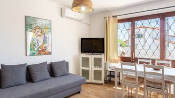 A living room with a gray sofa, a TV on a cabinet, a dining table with chairs, and a window with a view of buildings.