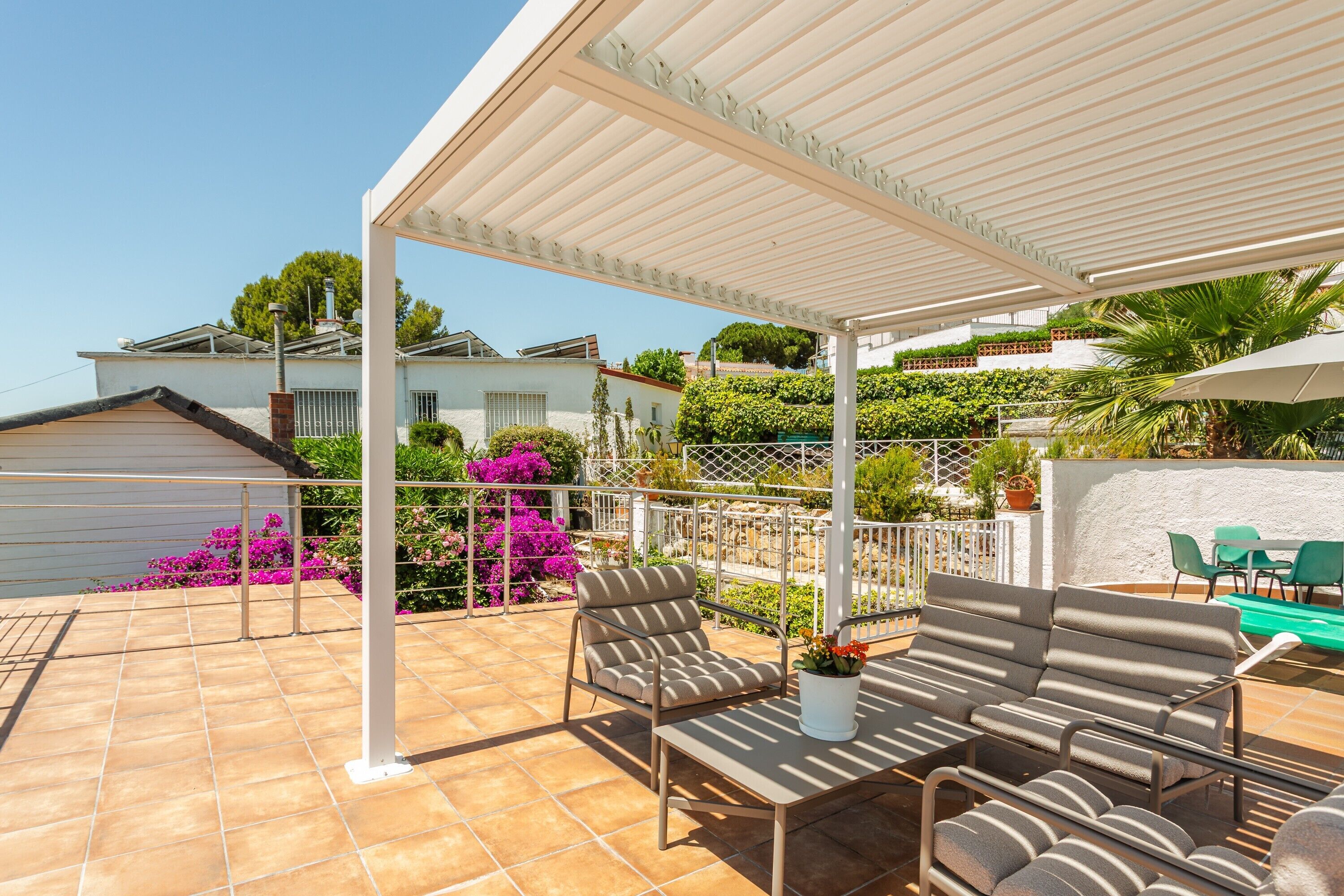 Eine überdachte Terrasse mit Gartenmöbeln, einer weißen Pergola und Blick auf Nachbarhäuser.