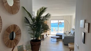 Un salón con una planta grande en maceta, un espejo redondo y vistas al mar desde un balcón.