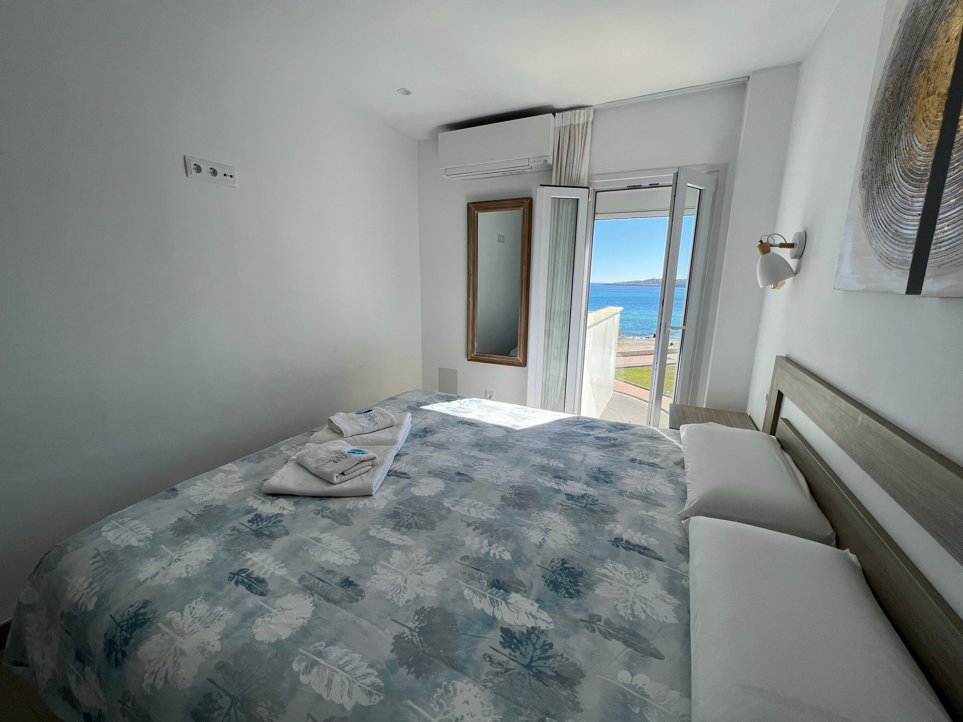 Una habitación de hotel con una cama, un ventanal con vistas al mar, una puerta y una lámpara fijada a la pared.