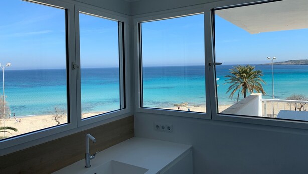 Una habitación con un ventanal grande con vista a una playa, un lavamanos blanco y un panel de madera en la pared.