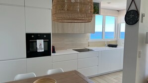 Una cocina moderna con armarios blancos, horno empotrado, lámpara colgante de mimbre y vistas al mar a través de grandes ventanales.