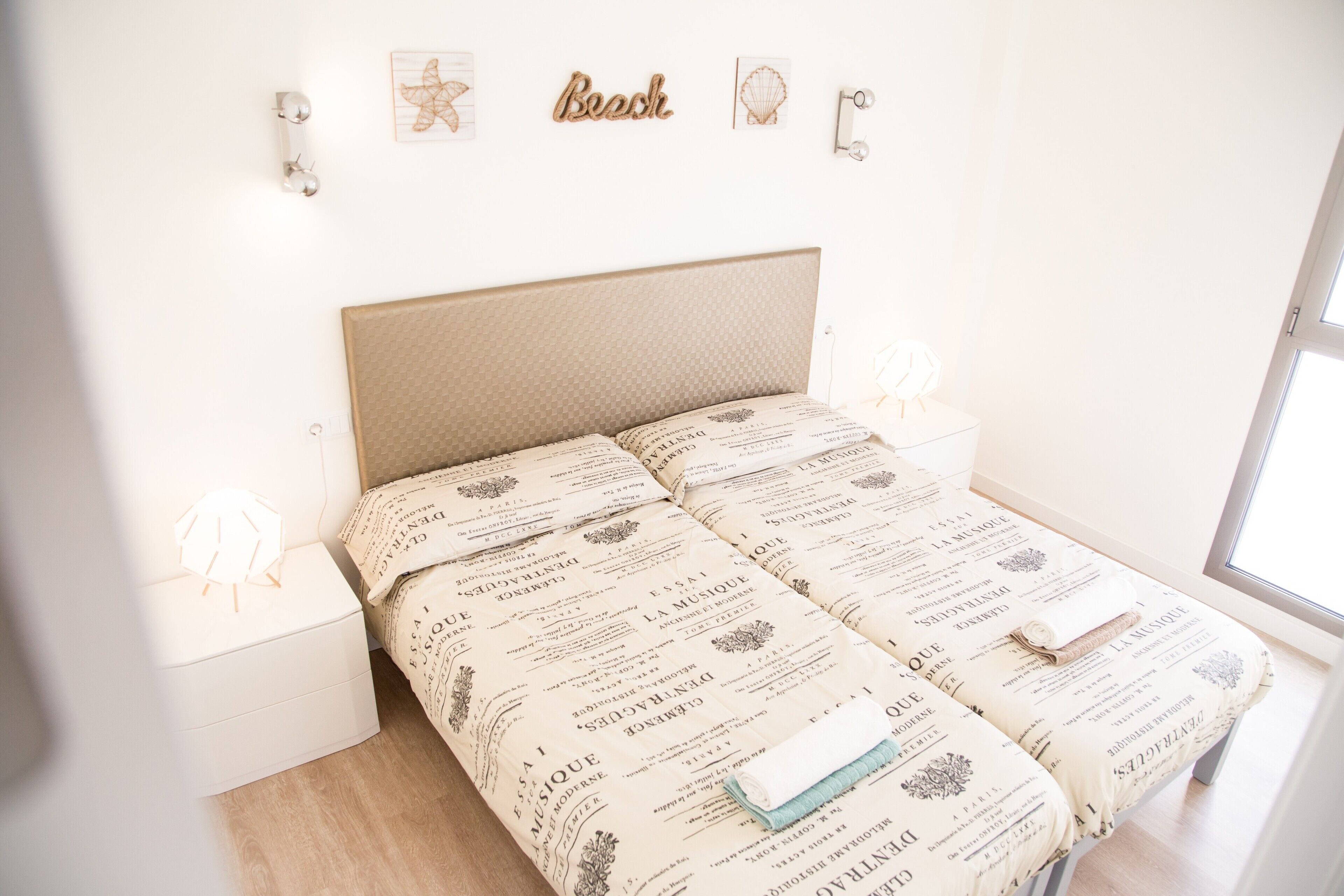 Una cama con cabecera color crema, mesitas de noche blancas y una pared decorada.