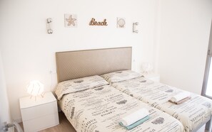 Une chambre à coucher avec un lit recouvert d’une couvre-lit à motifs, deux tables de chevet avec des lampes et des décorations murales comprenant une étoile de mer et un coquillage.