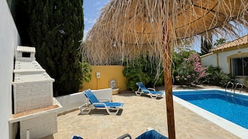 Un área de piscina con sillas de descanso, una parrilla y una sombrilla de paja.