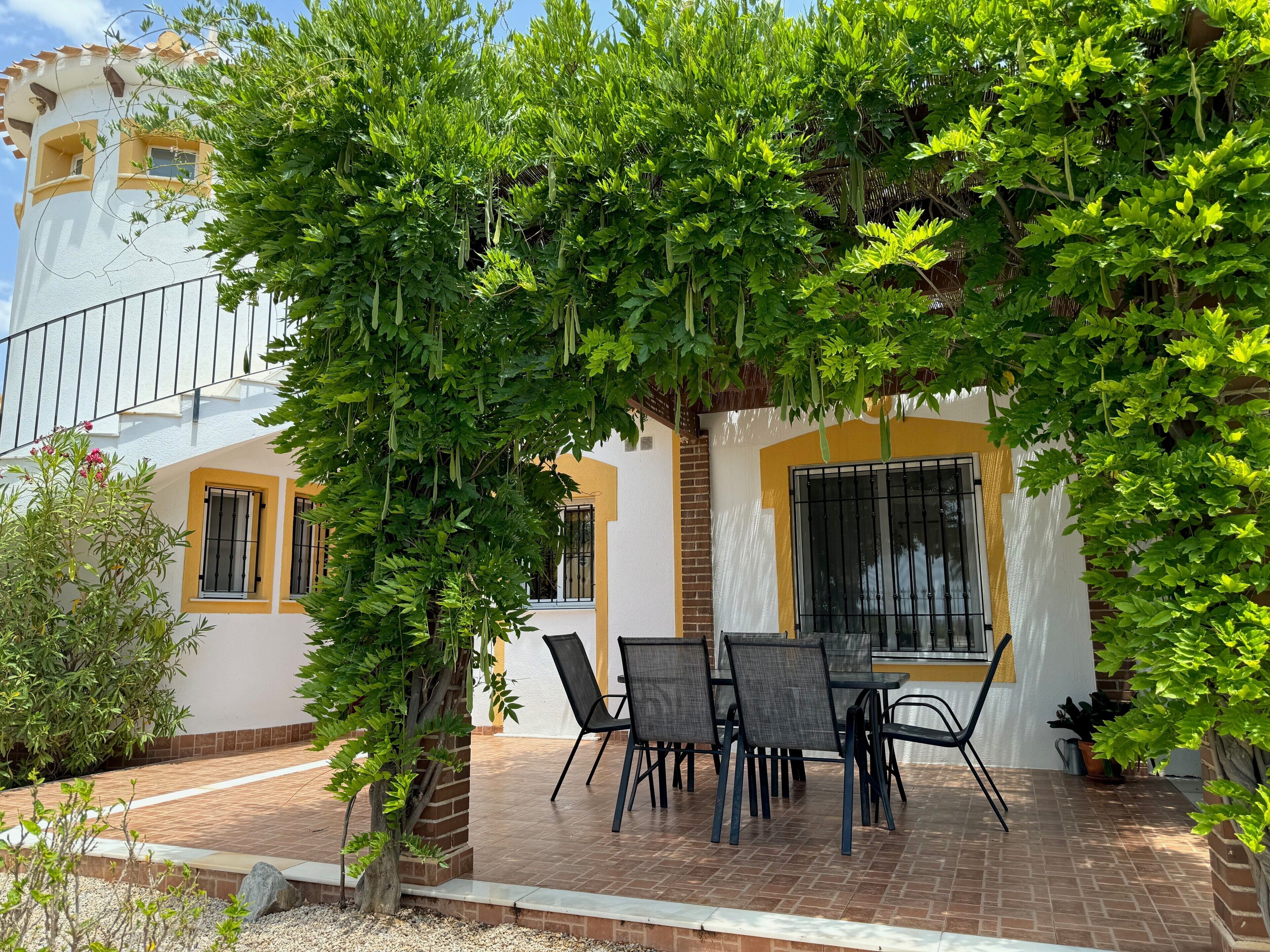 Un patio con mesa y sillas, rodeado de follaje y un edificio con paredes amarillas y blancas.