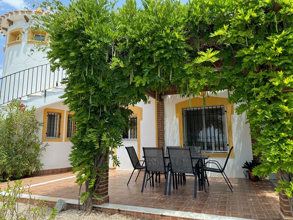 A patio with a table and chairs, surrounded by greenery and a building with yellow and white walls.