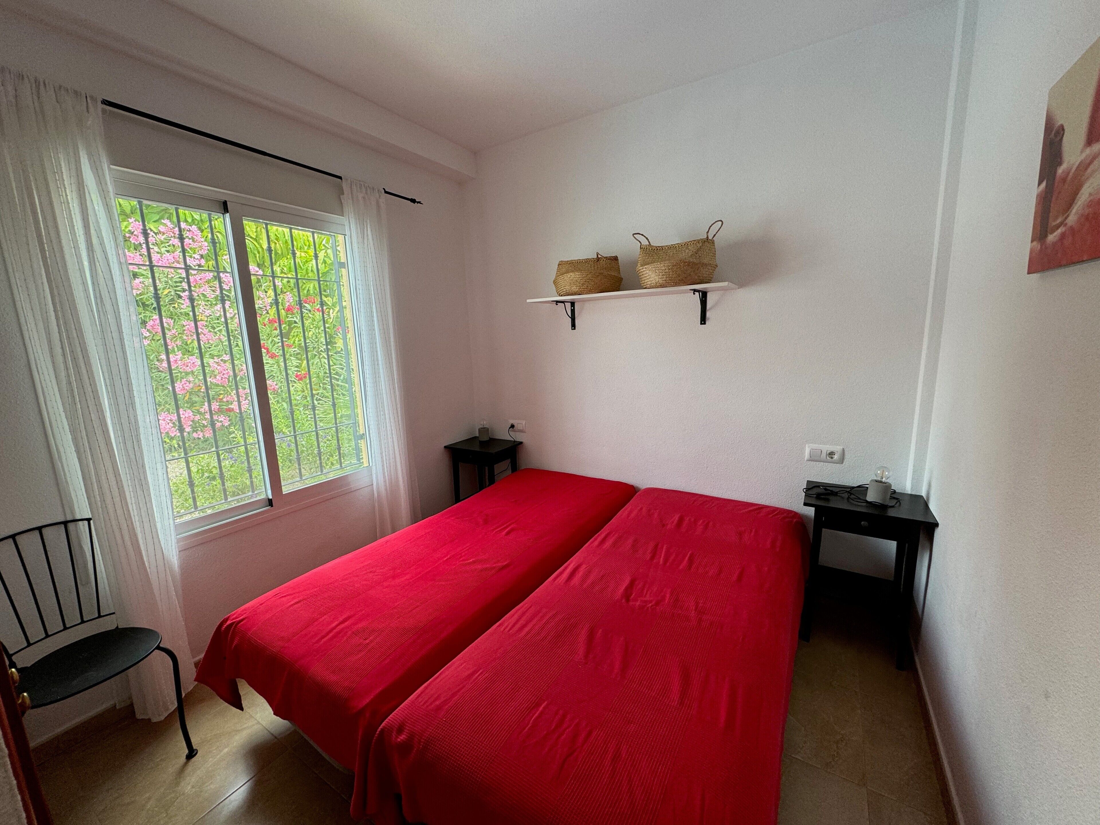 Una habitación con dos camas, una ventana con cortinas, una mesita pequeña y un estante con cestas.