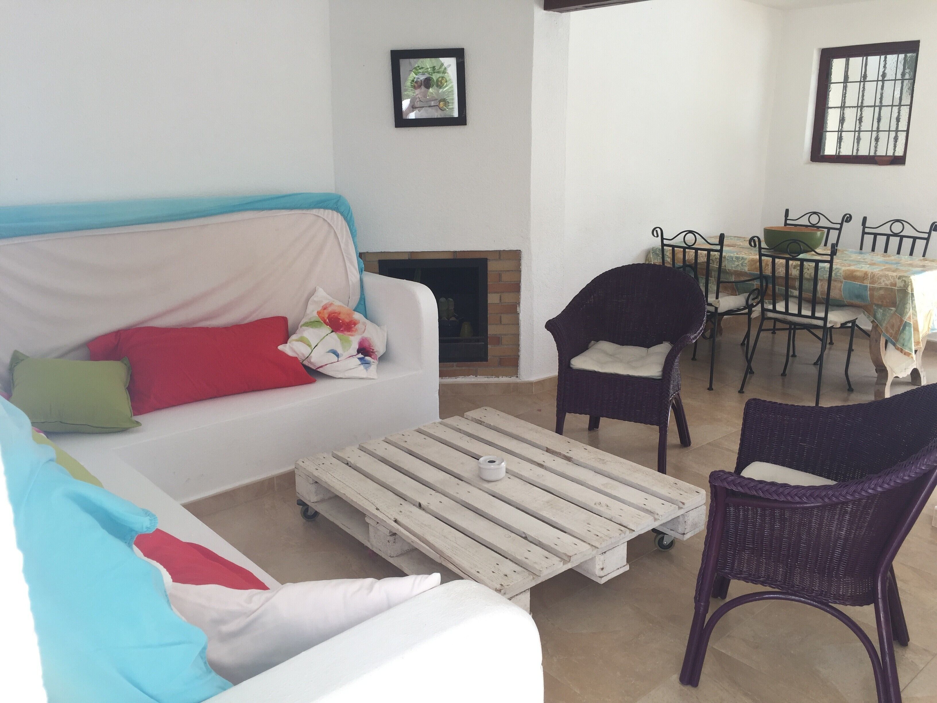 Un salon avec un canapé blanc, un fauteuil en osier, une table à manger avec des chaises et une cheminée.